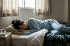 Person in light-blue pajamas sleeping on a bed in a softly lit bedroom, with a pillow under their head and a side table nearby