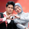 Couple on stage form a heart with their hands, the man in a red patterned jacket and white shirt, the woman in a light blue hijab and silver dress smile softly against a warm, blurred background.