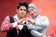 Couple on stage form a heart with their hands, the man in a red patterned jacket and white shirt, the woman in a light blue hijab and silver dress smile softly against a warm, blurred background.