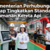 Inside a train maintenance workshop: officials in helmets beside a touchscreen control kiosk with a red train in the background, conveying safety standard upgrades by the Ministry of Transport (Indonesia).