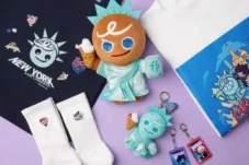 Plush Statue of Liberty–inspired toy with ice cream cone, surrounded by white socks, a navy tote bag, and colorful keychains on a purple background.