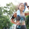 Woman in workout clothes wiping sweat with towel after running outdoors, holding a water bottle and wearing an armband with phone.