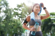Woman in workout clothes wiping sweat with towel after running outdoors, holding a water bottle and wearing an armband with phone.