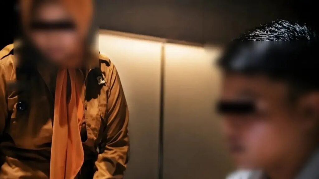 Two men in an elevator with their eyes obscured by black bars; one wears a brown shirt with a light pink tie, the other is blurred in the foreground.
