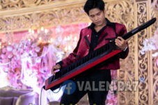 Male musician in a maroon jacket playing a red keytar on an ornate, flower-decorated stage backdrop.
