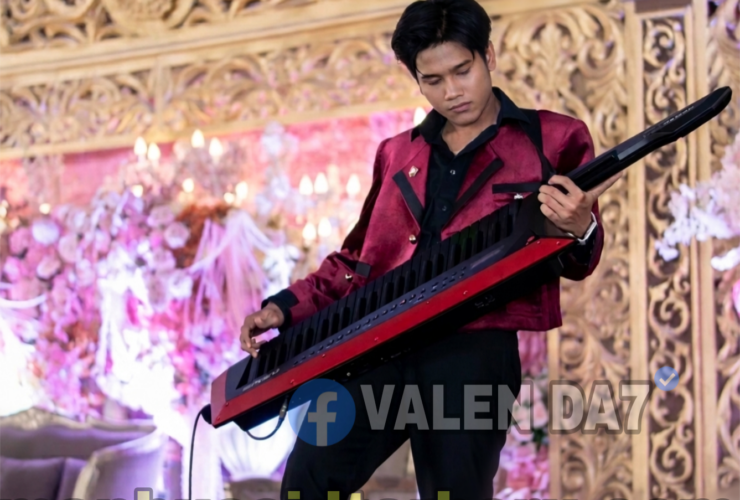 Male musician in a maroon jacket playing a red keytar on an ornate, flower-decorated stage backdrop.