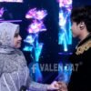 Two performers on a stage hold hands and smile at each other, dressed in elaborate silver and gold outfits with a colorful LED backdrop.