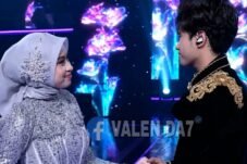 Two performers on a stage hold hands and smile at each other, dressed in elaborate silver and gold outfits with a colorful LED backdrop.