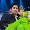 Young man in a black blazer and neon green pants sits in a backstage area, waving at the camera with blue stage lights behind him.