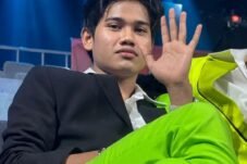 Young man in a black blazer and neon green pants sits in a backstage area, waving at the camera with blue stage lights behind him.
