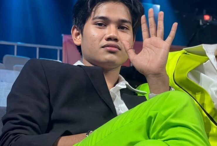 Young man in a black blazer and neon green pants sits in a backstage area, waving at the camera with blue stage lights behind him.