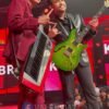 Two male musicians on a red-lit stage pose with a keyboard (left) and a bright green electric guitar (right).