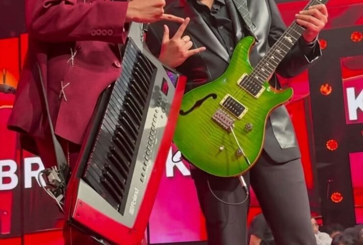 Two male musicians on a red-lit stage pose with a keyboard (left) and a bright green electric guitar (right).